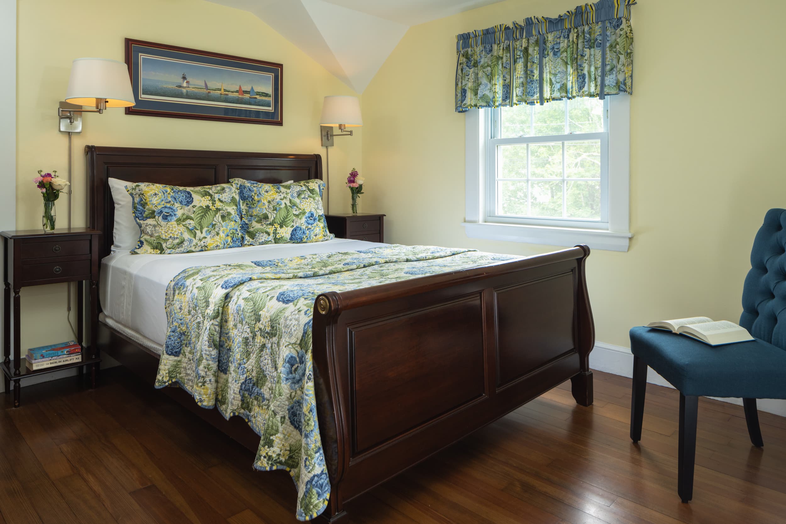 A peaceful bedroom with light yellow walls and a dark wood queen bed. The bed is dressed with a blue and yellow floral comforter, and the room has two bedside lamps and a window that lets in natural sunlight.