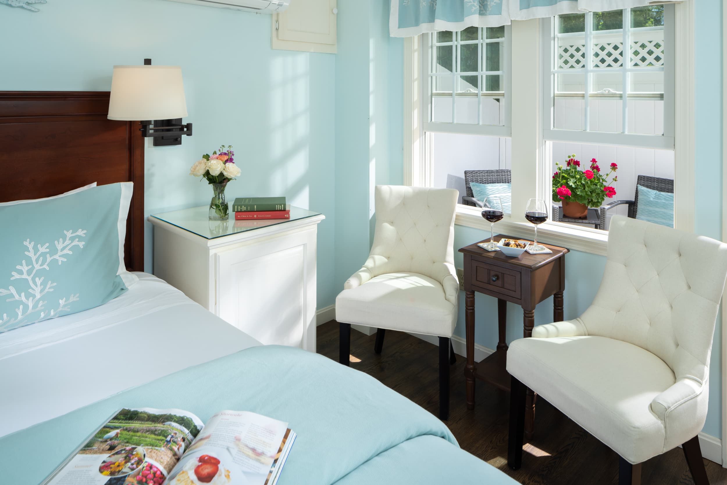 A peaceful bedroom with light blue walls and a dark wood bed frame. The room has two white tufted chairs and a small table with two glasses of red wine, offering a perfect spot to relax by the window.