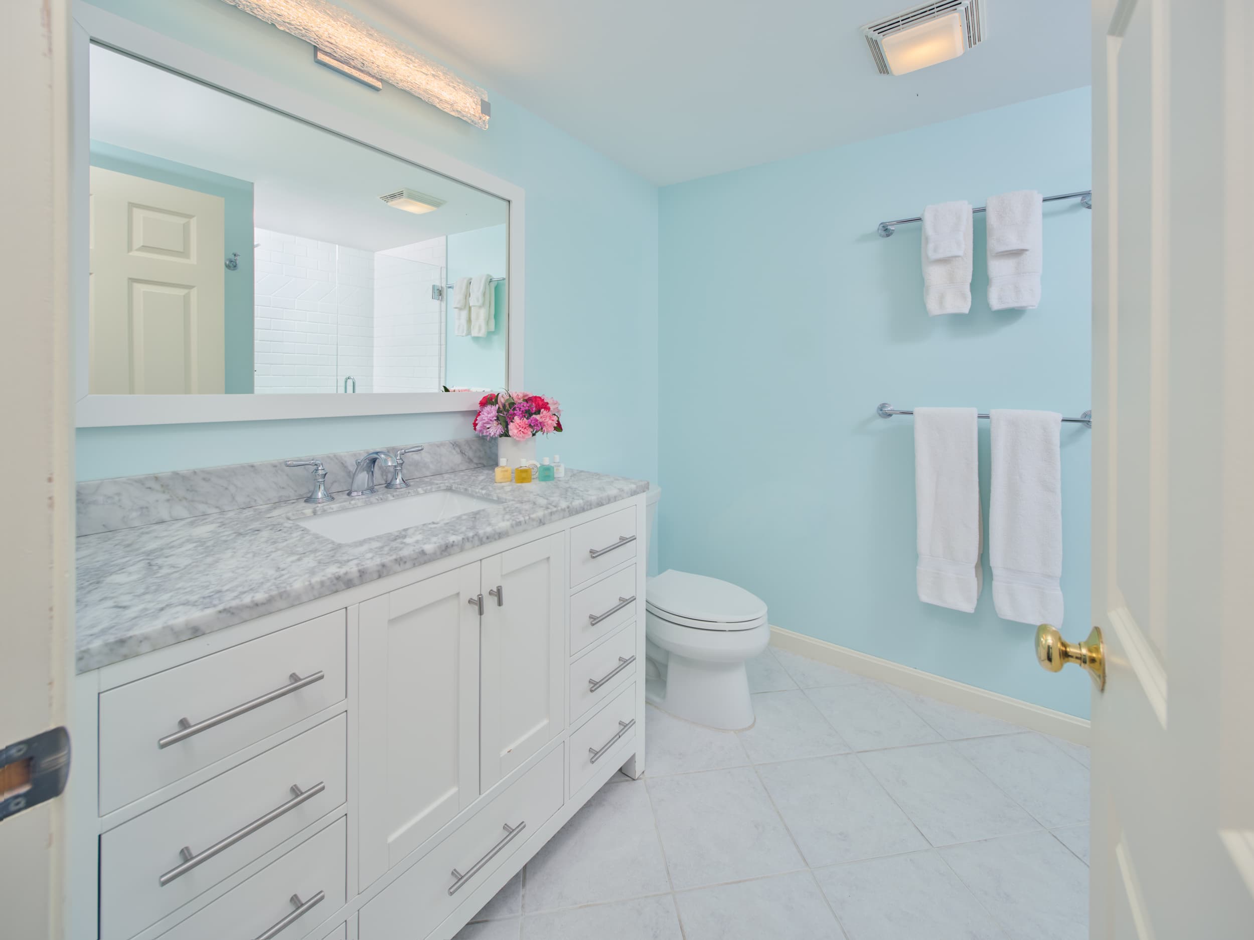 A modern bathroom with light blue walls, white toilet, step-in shower and a white vanity with a marble countertop. A bouquet of hydrangeas and other flowers is on the counter, and fresh white towels are ready for use.