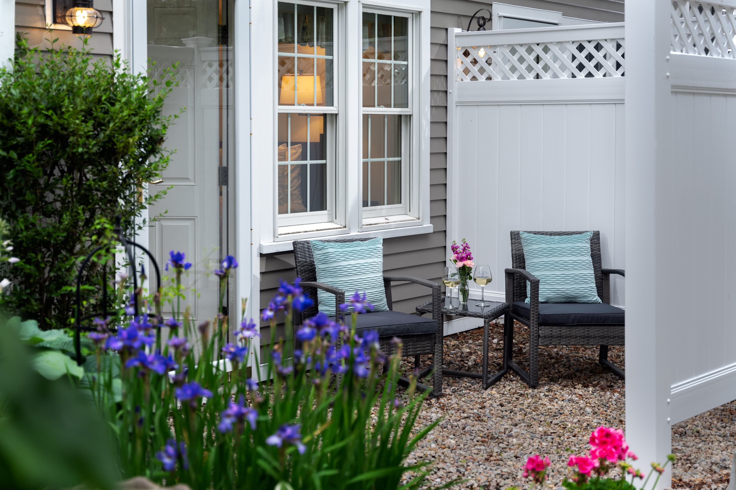 A peaceful outdoor patio area with two chairs and a small table. The chairs have blue pillows, and a small flower bouquet is on the table. A white lattice fence separates the space from the surroundings.