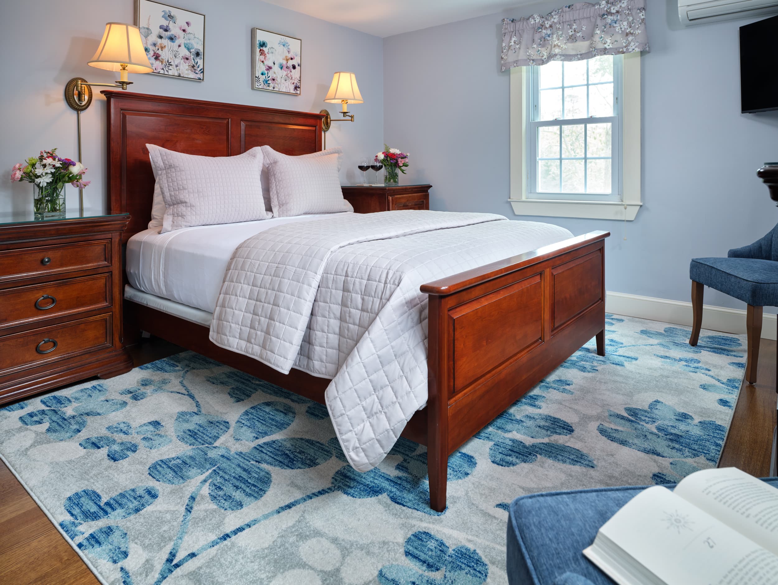 A comfortable bedroom with light blue walls and a dark wood bed frame. The room has a floral blue and white rug and a matching blue armchair.