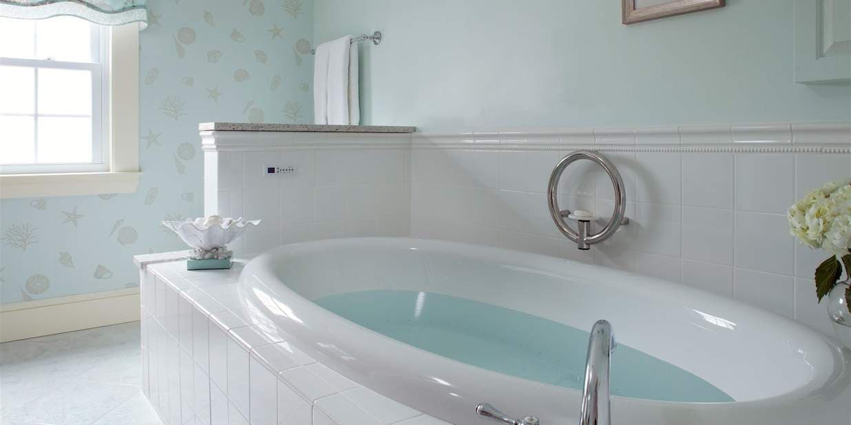 A light and airy bathroom with a large, white soaking tub filled with water. The room features white tile, light-blue walls, and a window for natural light.