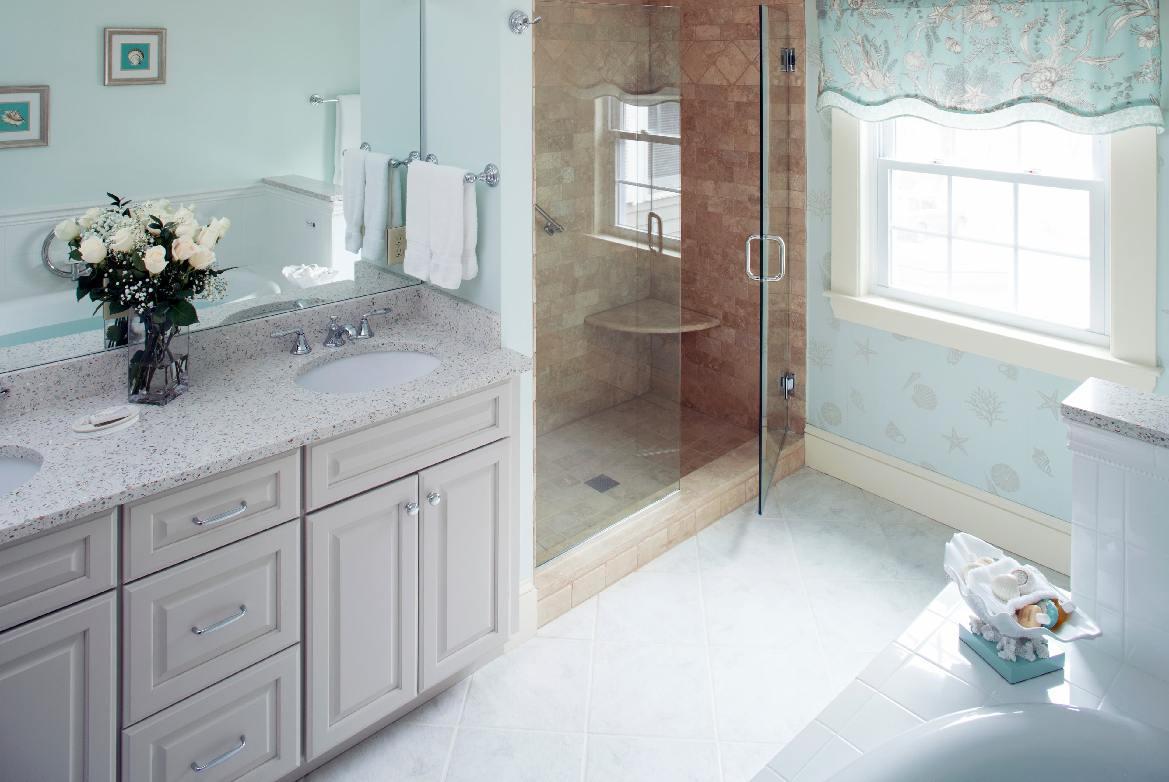 A large, elegant bathroom with a double vanity and a glass shower. The room has light-blue decor, a window letting in natural light, and a bouquet of white roses on the counter.