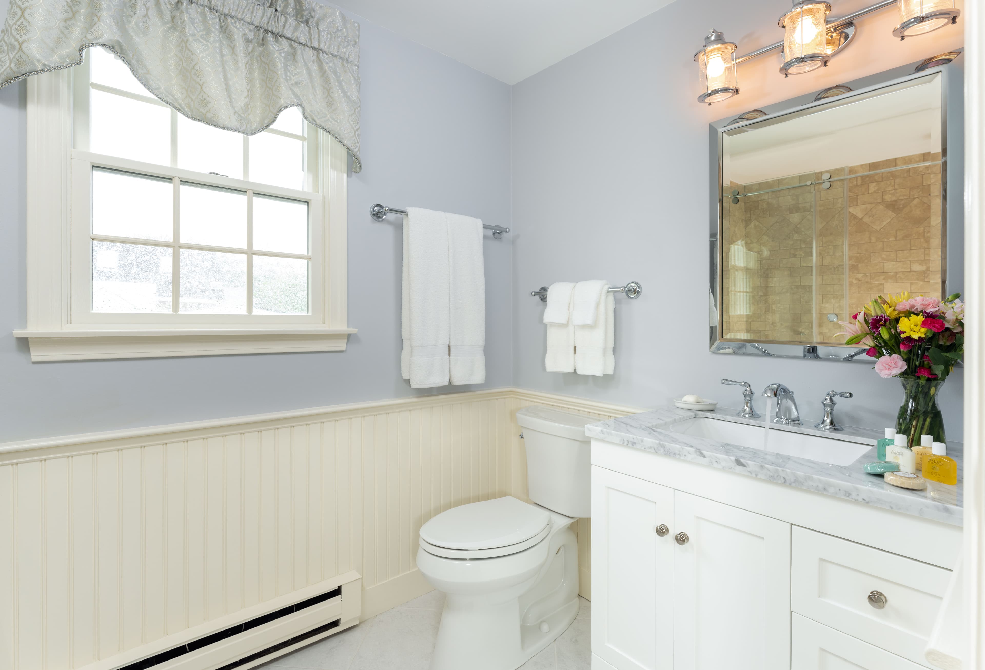 A cozy, bright bathroom with light-blue walls and white wainscoting. A vanity with a marble countertop and a mirror is on the right, and a window is on the left.
