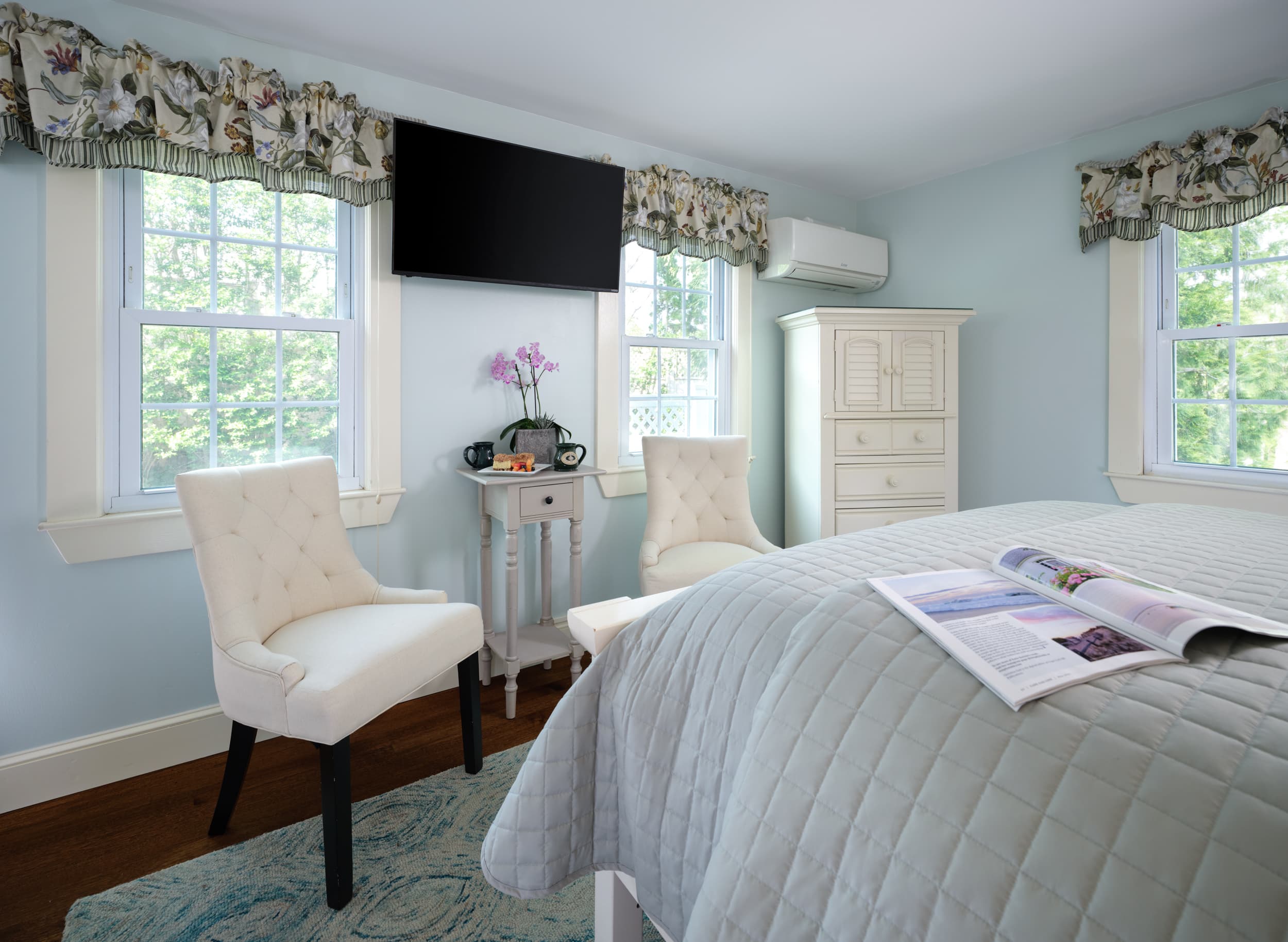 A tranquil bedroom with soft blue walls and two tufted chairs. The room has a queen bed with light blue bedding, a flat-screen TV mounted on the wall, and multiple large windows that bring in natural light.