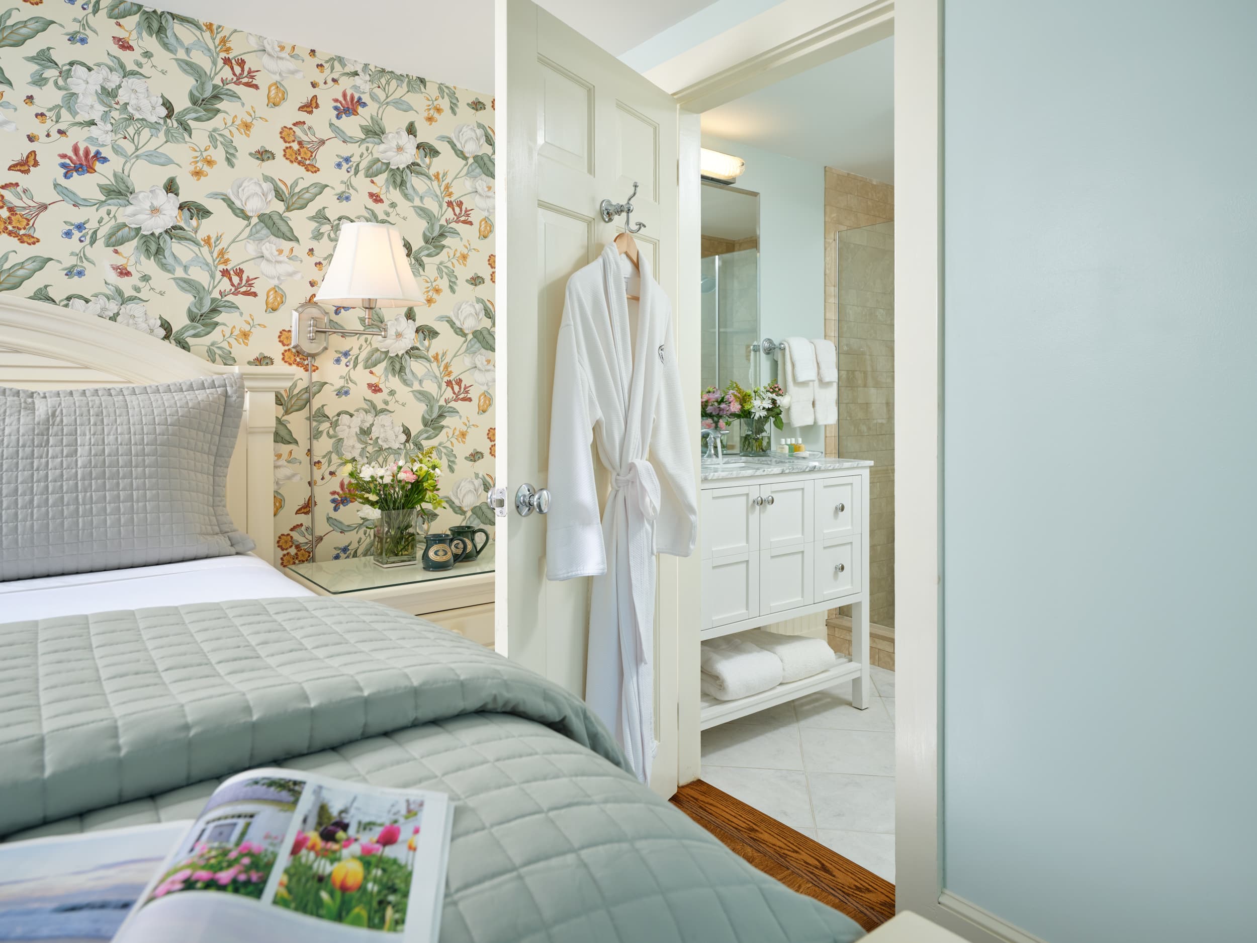 A tranquil bedroom with an open door to an ensuite bathroom. The bedroom has a queen bed with a light blue comforter and pillows, while the bathroom has a white vanity and a robe hanging on the door.