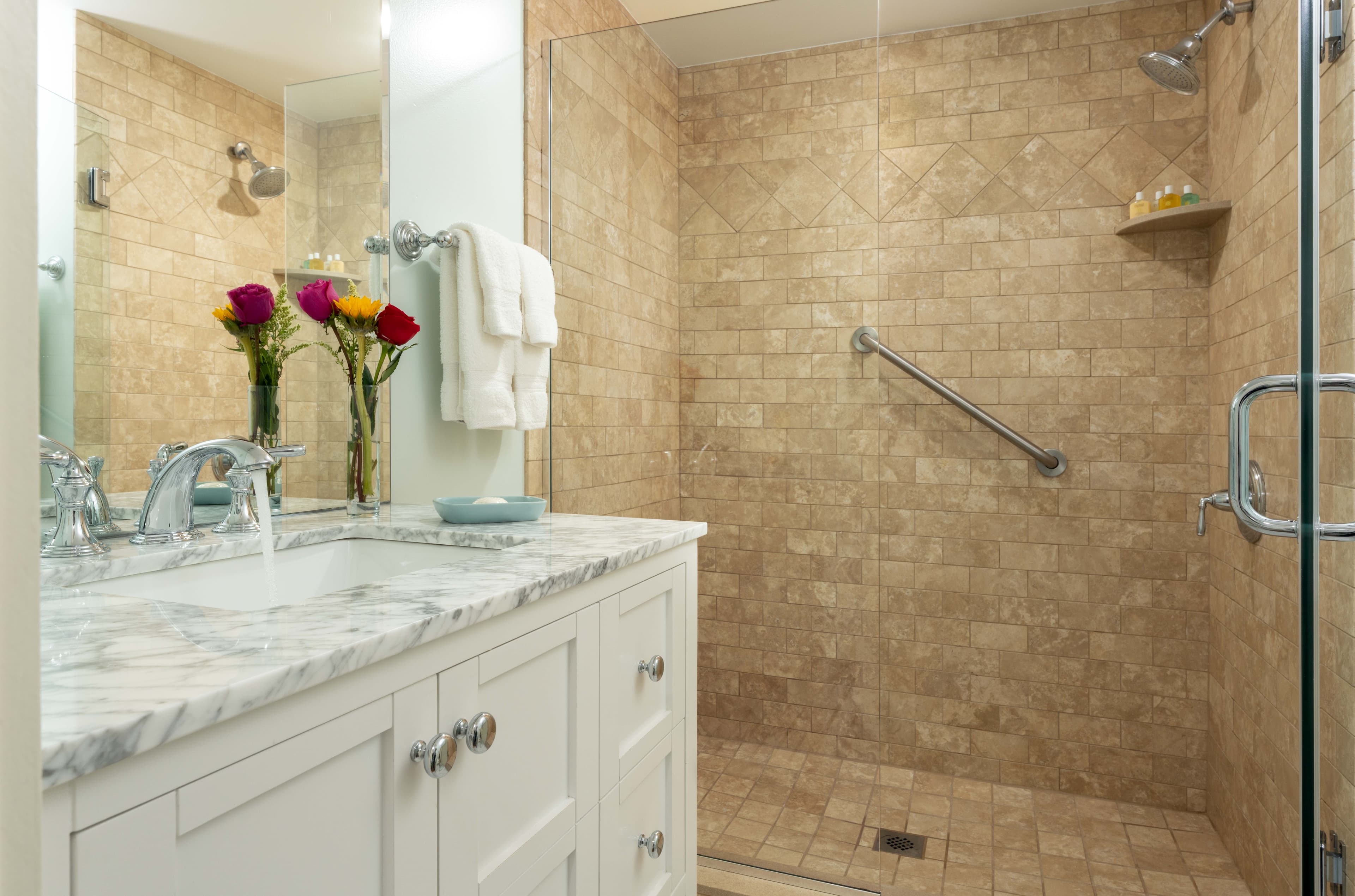 A spacious bathroom with a large vanity and a glass walk-in shower. The shower is tiled in a light stone, with a metal grab bar and a built-in corner shelf.
