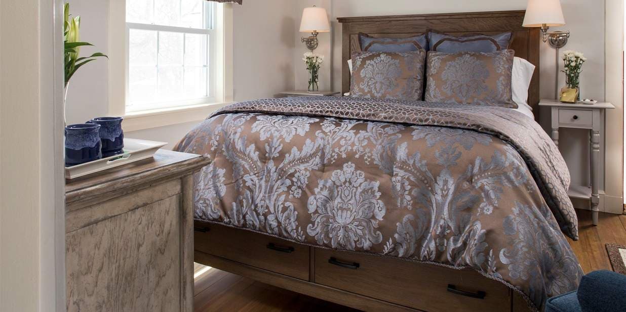 A bedroom with a rustic wood queen bed and a silver comforter with a textured pattern. The bed has built-in storage drawers and is flanked by matching nightstands with lamps.