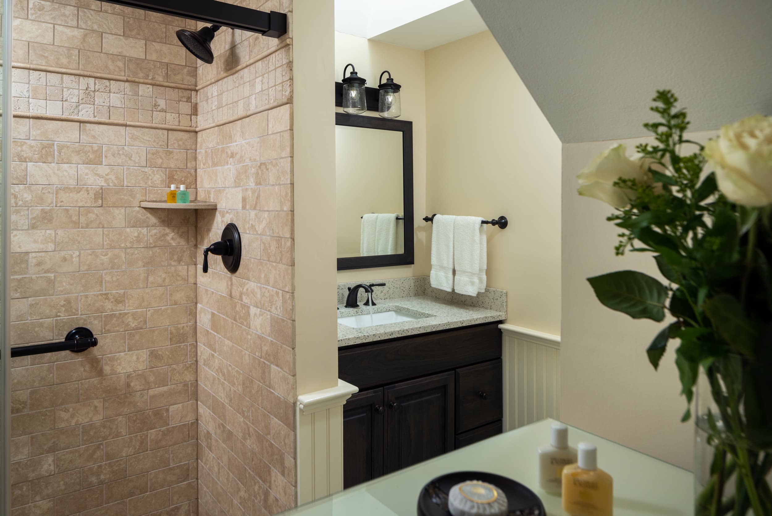 A modern bathroom with a walk-in shower. The shower walls are tiled in a light, natural stone with dark bronze fixtures. A single vanity with a white top is visible, and a bouquet of white roses sits on a glass countertop in the foreground.