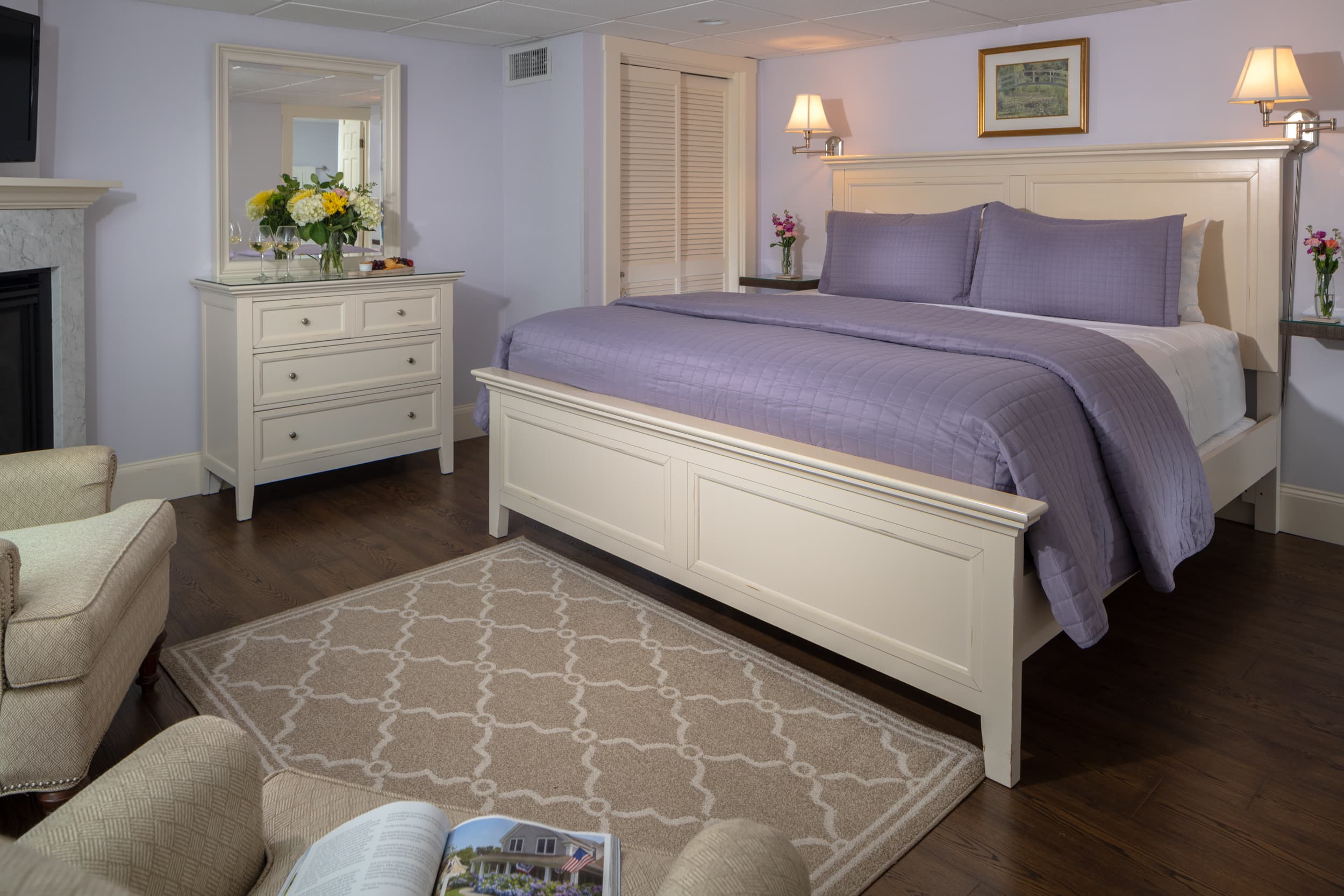 A serene bedroom with light purple walls and a white-painted wooden bed frame. A bouquet of flowers sits on a dresser, and a fireplace is on the left.