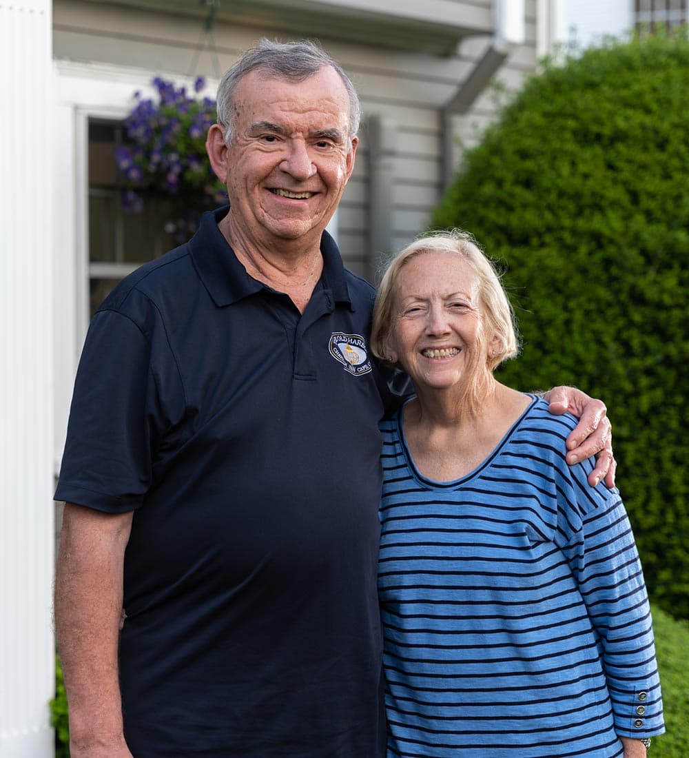 An older man and woman smile together outside a house, with the man wearing a black polo shirt and the woman in a striped blue top.