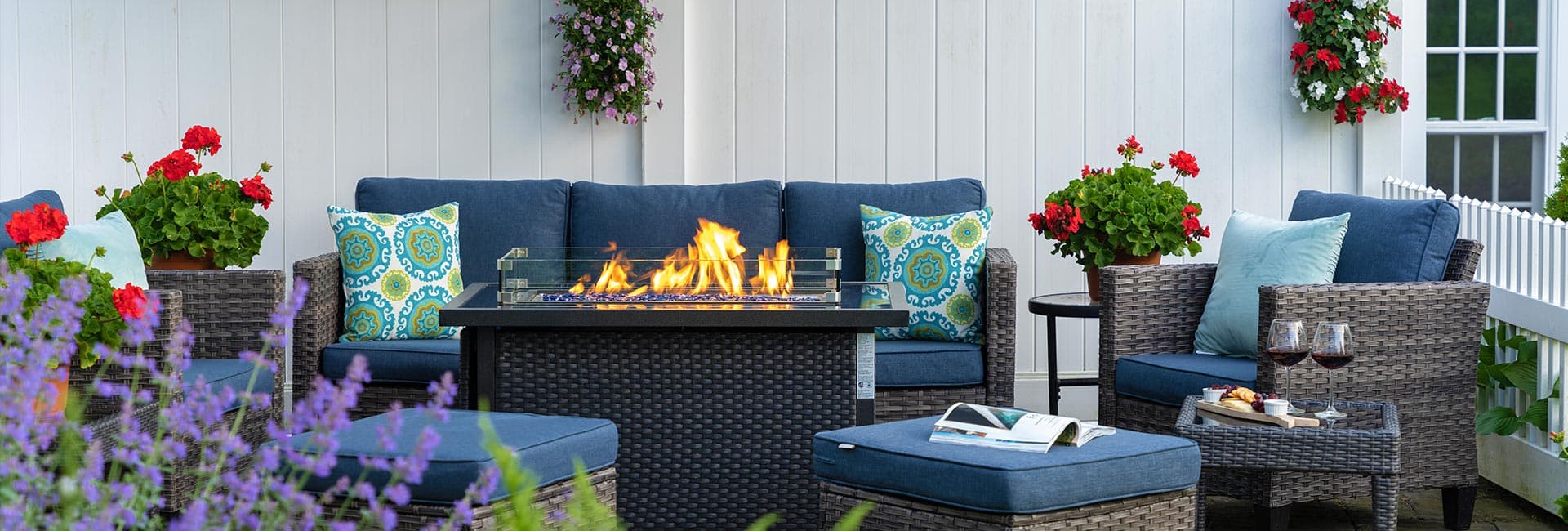 A cozy outdoor seating area featuring blue cushions, flowering plants, and a fire pit.