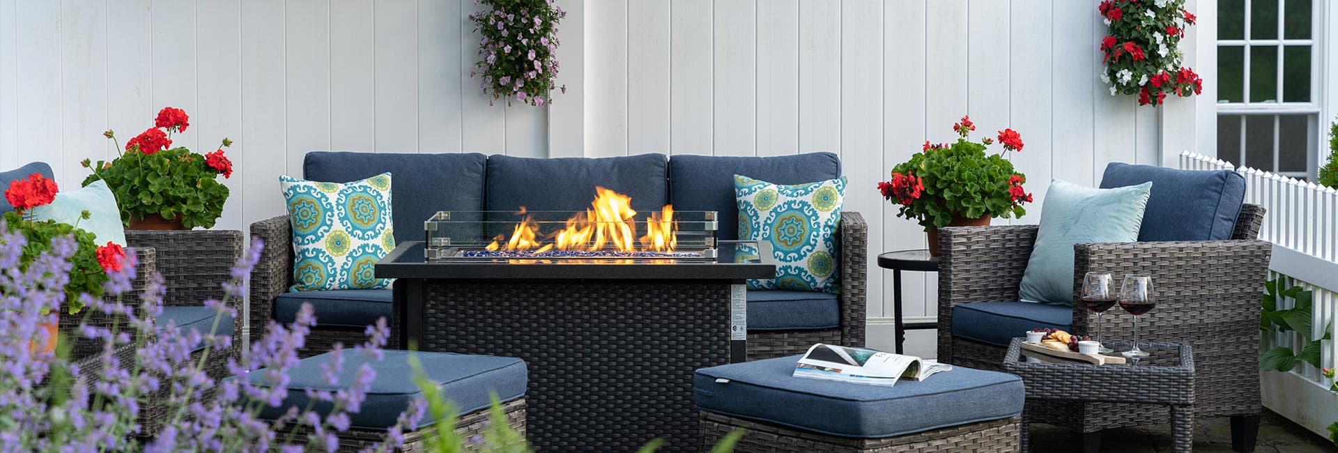 A cozy outdoor seating area with a fire pit, surrounded by flowering plants and decorative pillows.