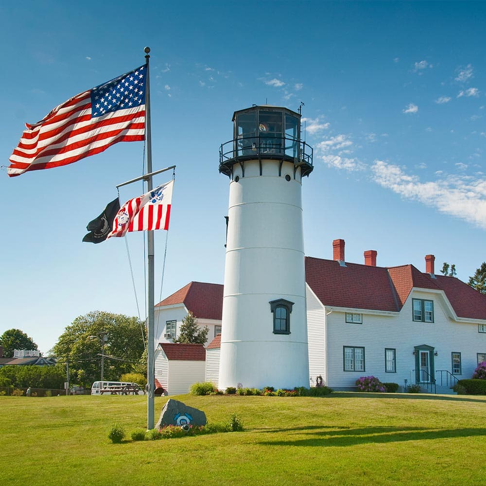 A white lighthouse with a light beacon stands beside a house, with American flags flying in front.