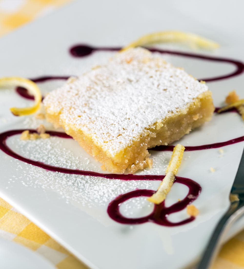 A lemon bar dusted with powdered sugar, served with berry sauce and lemon zest.