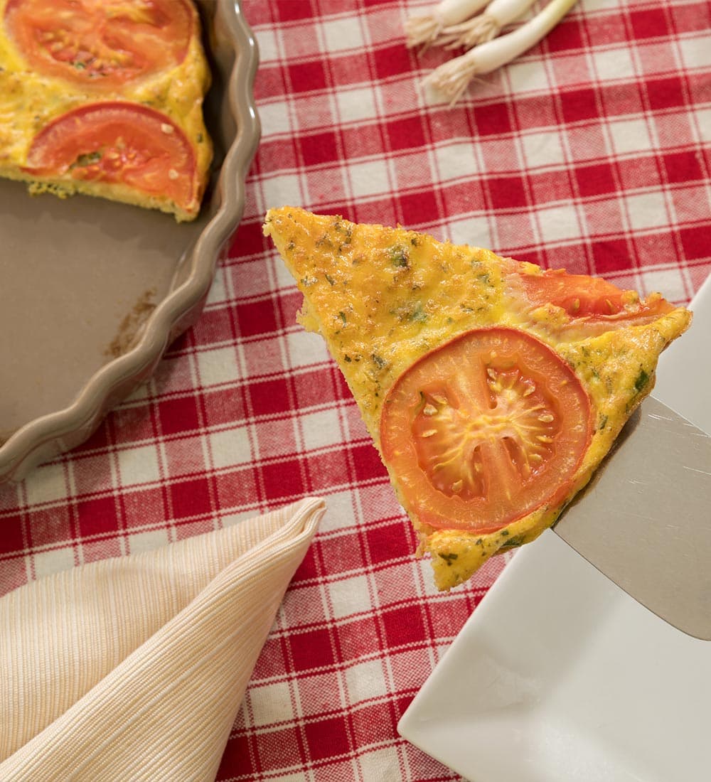 A slice of tomato frittata being lifted from a pie dish onto a white plate, on a red and white checkered tablecloth.