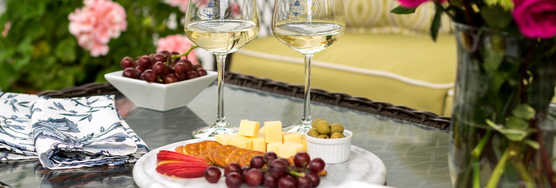 A table setting featuring white wine glasses, a platter of cheese, crackers, fruits, and olives, surrounded by flowers.