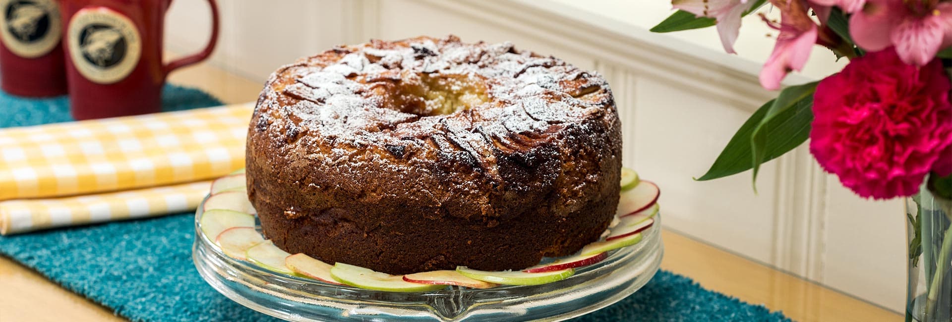A round cake dusted with powdered sugar, displayed on a glass plate with apple slices and surrounded by flowers and cups.