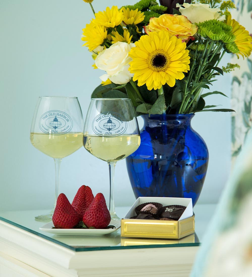 Two glasses of white wine are placed beside a vibrant bouquet of flowers, accompanied by fresh strawberries and a box of chocolates on a glass table.
