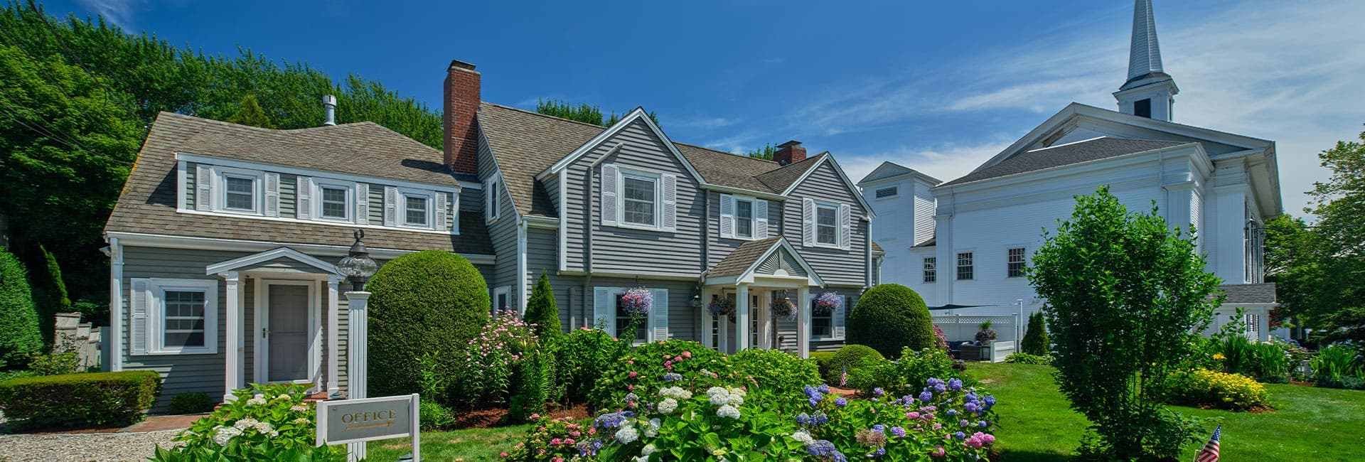 A charming two-story gray house with lush gardens and a white church spire nearby.