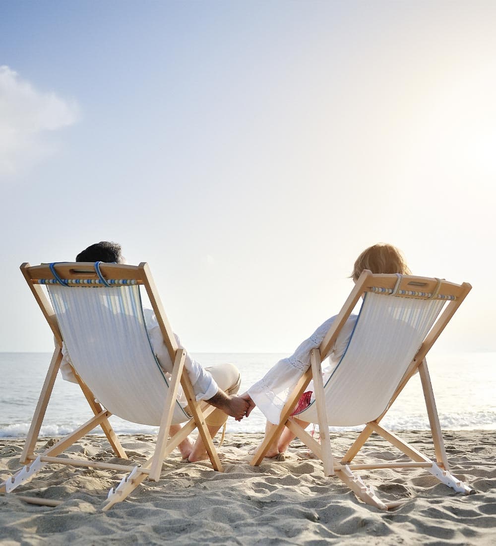 A couple sits in beach chairs holding hands, gazing at the ocean.