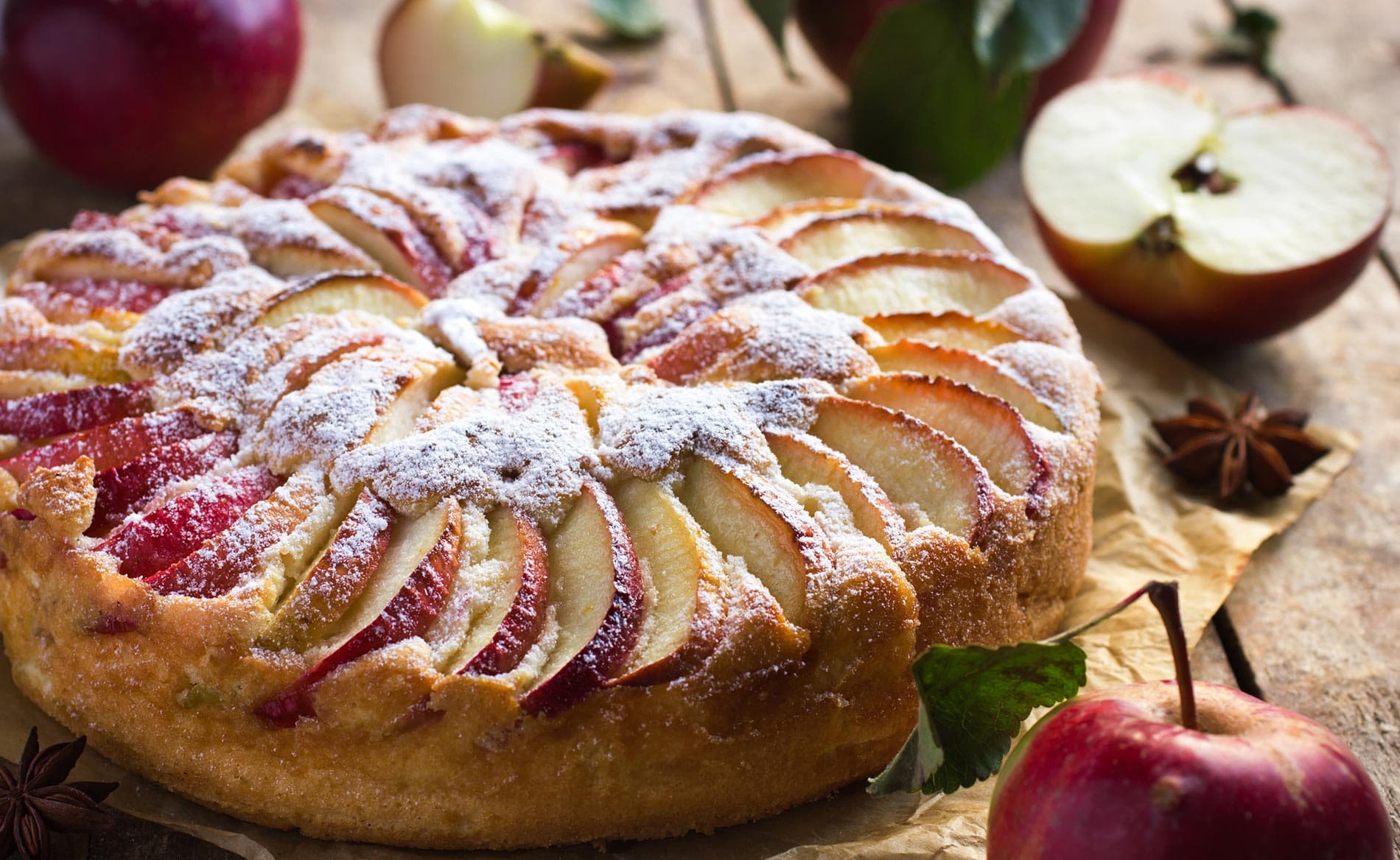 A freshly baked apple tart dusted with powdered sugar, surrounded by apples and greenery on a wooden surface. A freshly baked apple tart dusted with powdered sugar, surrounded by apples and greenery on a wooden surface.