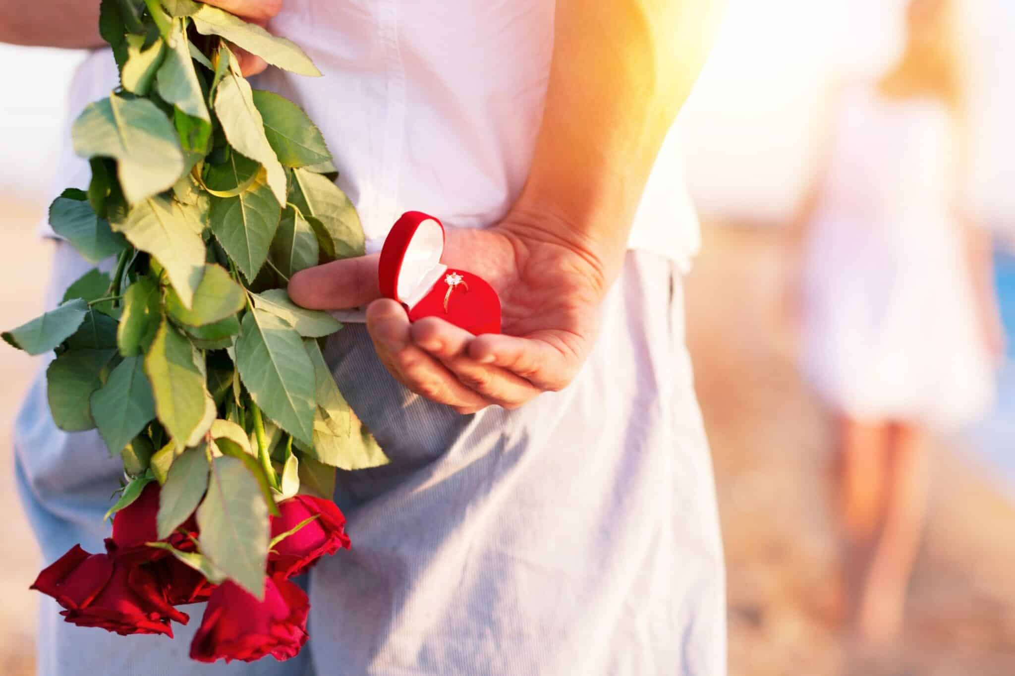A person holds a bouquet of roses and an open ring box behind their back while a figure walks away in the background. A person holds a bouquet of roses and an open ring box behind their back while a figure walks away in the background.