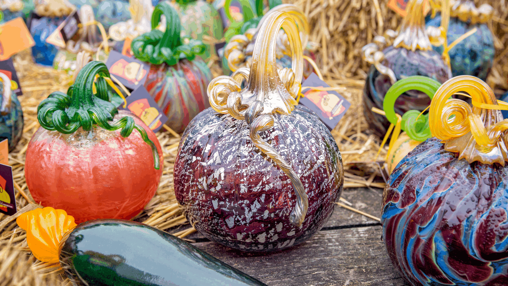 A collection of colorful glass pumpkins displayed on straw with decorative tags. A collection of colorful glass pumpkins displayed on straw with decorative tags.