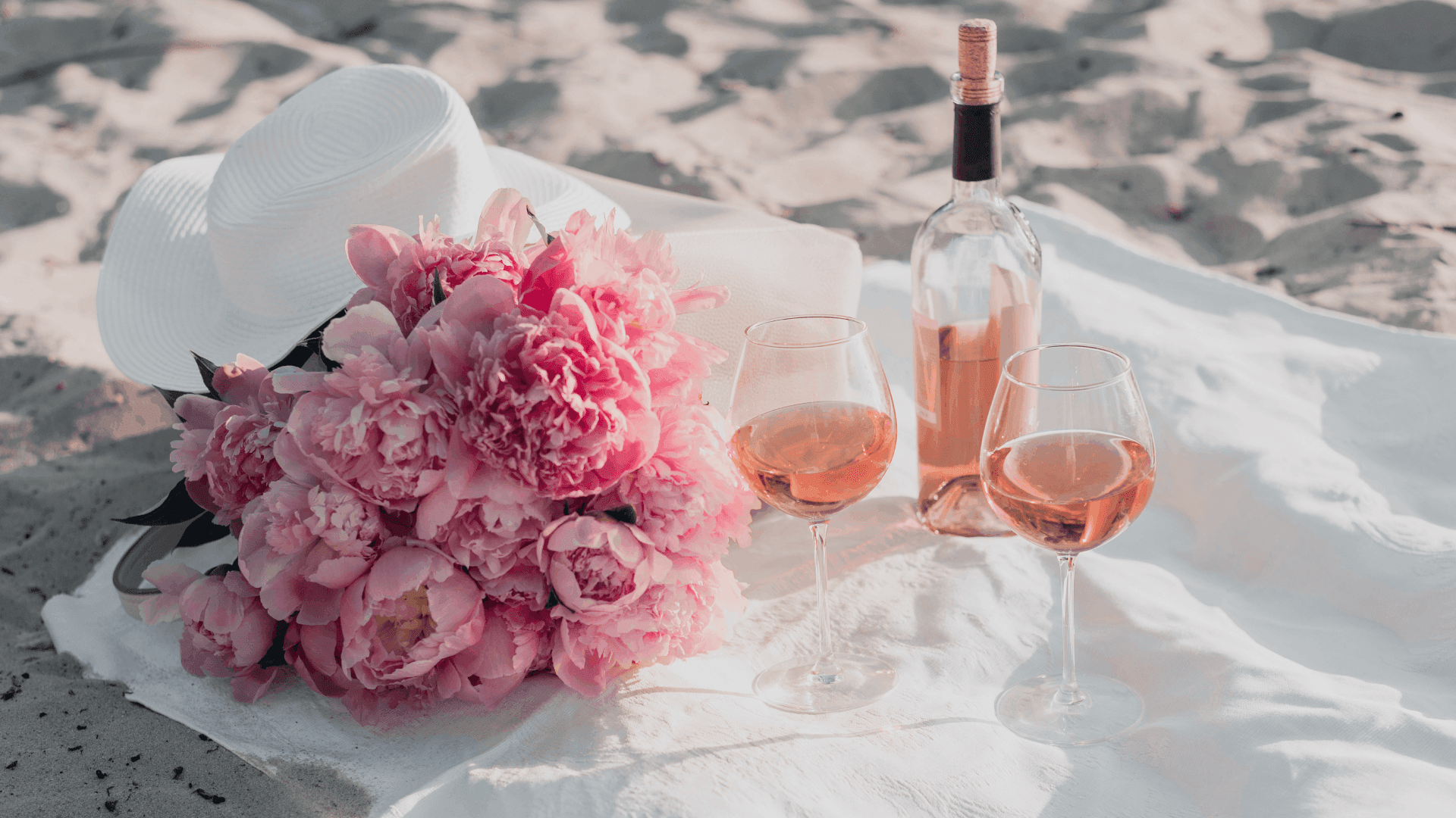 A bouquet of pink peonies, two glasses of rosé, and a bottle of wine are arranged on a white cloth in a sandy setting. A bouquet of pink peonies, two glasses of rosé, and a bottle of wine are arranged on a white cloth in a sandy setting.