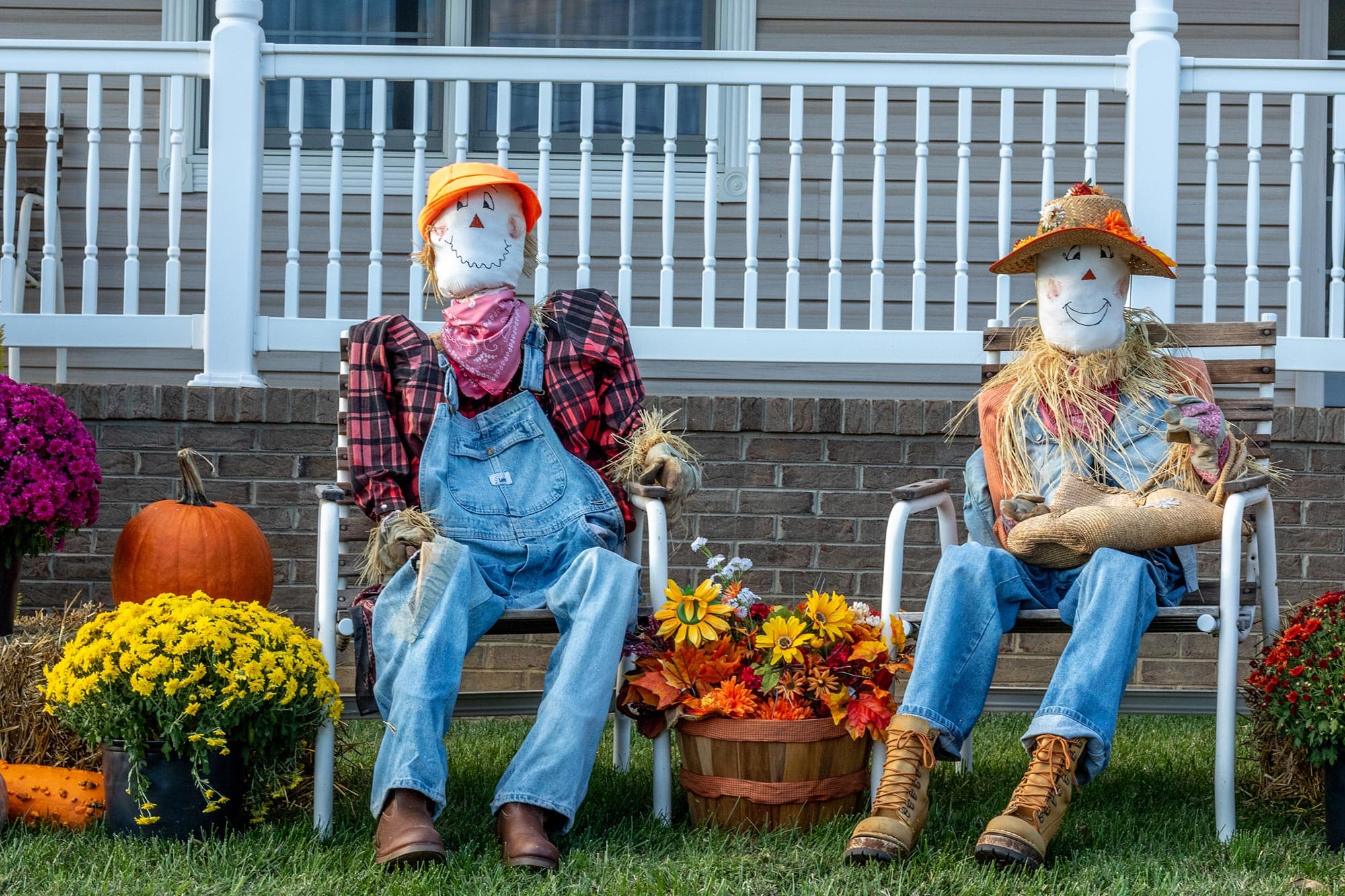 Two scarecrows dressed in colorful outfits sit on chairs surrounded by autumn flowers and a pumpkin. Two scarecrows dressed in colorful outfits sit on chairs surrounded by autumn flowers and a pumpkin.